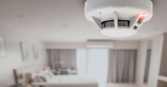 A close up of a smoke detector on the ceiling of a white room. There's furniture and a window in the background.
